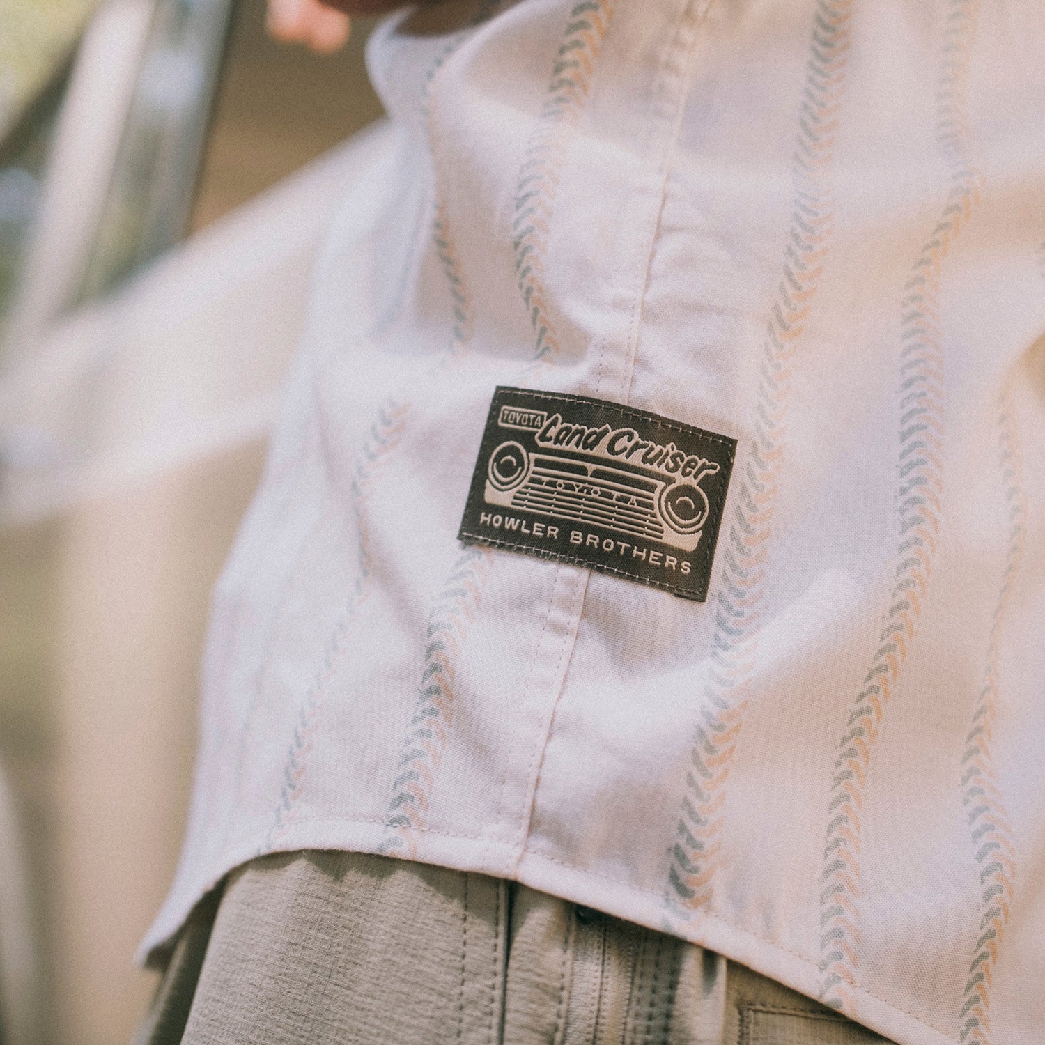 A close-up of a vintage inspired Howler Brothers pearl snap shirt, white with blue stripes and a patch reading Land Cruiser Howler Brothers with a simple car graphic, worn outdoors. - View 6