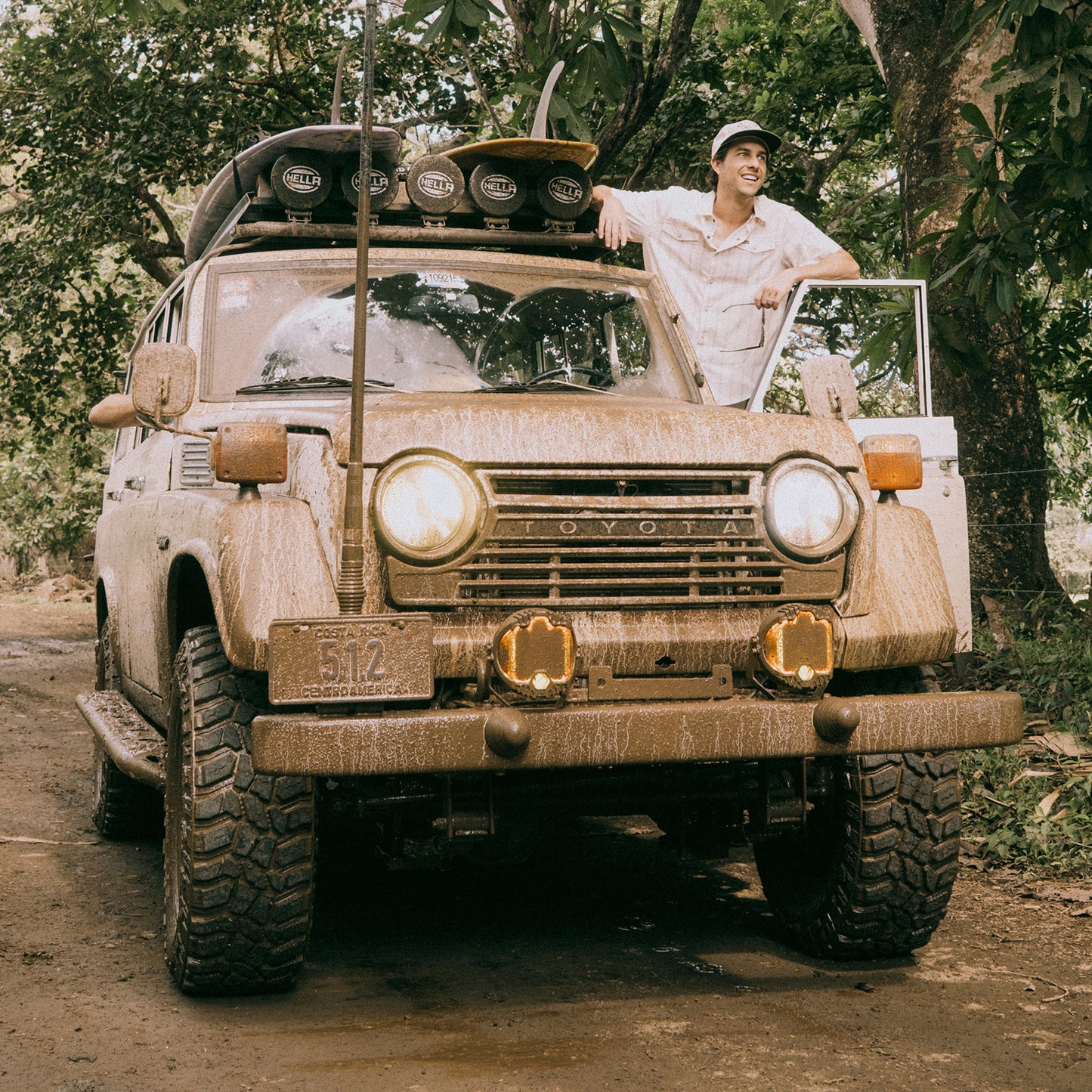 A person in a Howler Brothers pearl snap shirt and cap stands smiling next to an old, mud-covered Toyota SUV with surfboards on the roof, parked on a dirt road surrounded by green trees. - View 7