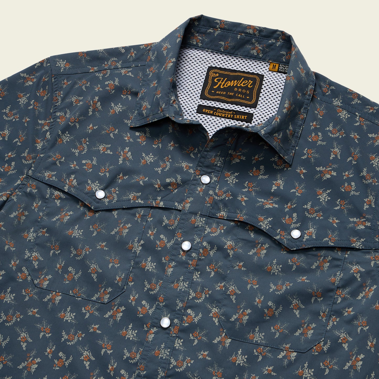 A close-up of a blue pearl snap shirt with a small floral pattern, pointed chest pockets, vented rear yoke, and a visible “Howler” brand label inside the collar. - View 3