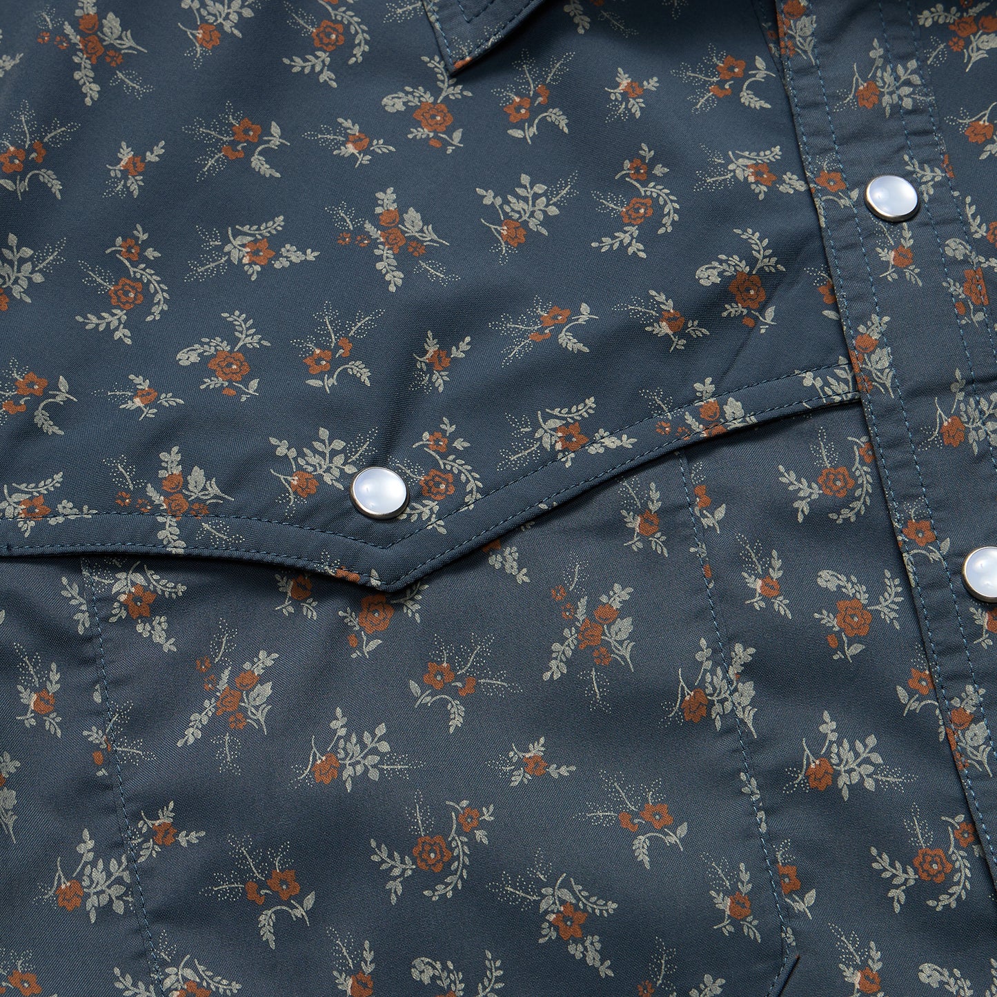 Close-up of a dark blue pearl snap shirt with a small floral pattern of red and beige flowers and green leaves. The fabric has white snaps, visible stitching, and suggests part of a technical utility shirt pocket. - View 6