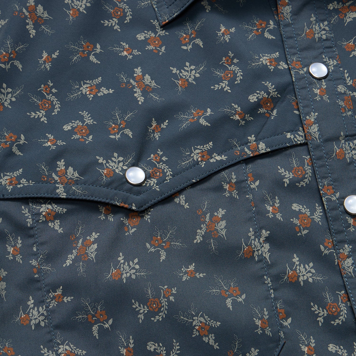 Close-up of a dark blue pearl snap shirt with a small floral pattern of red and beige flowers and green leaves. The fabric has white snaps, visible stitching, and suggests part of a technical utility shirt pocket. - View 6