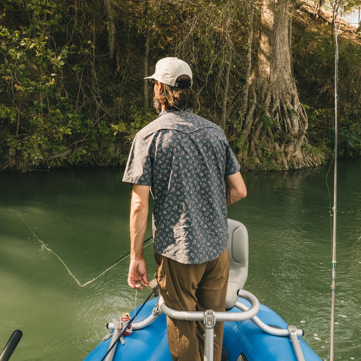 A man in a pearl snap shirt and cap stands at the edge of a blue inflatable boat, fishing on a calm river surrounded by lush greenery and tree roots along the riverbank. - View 8