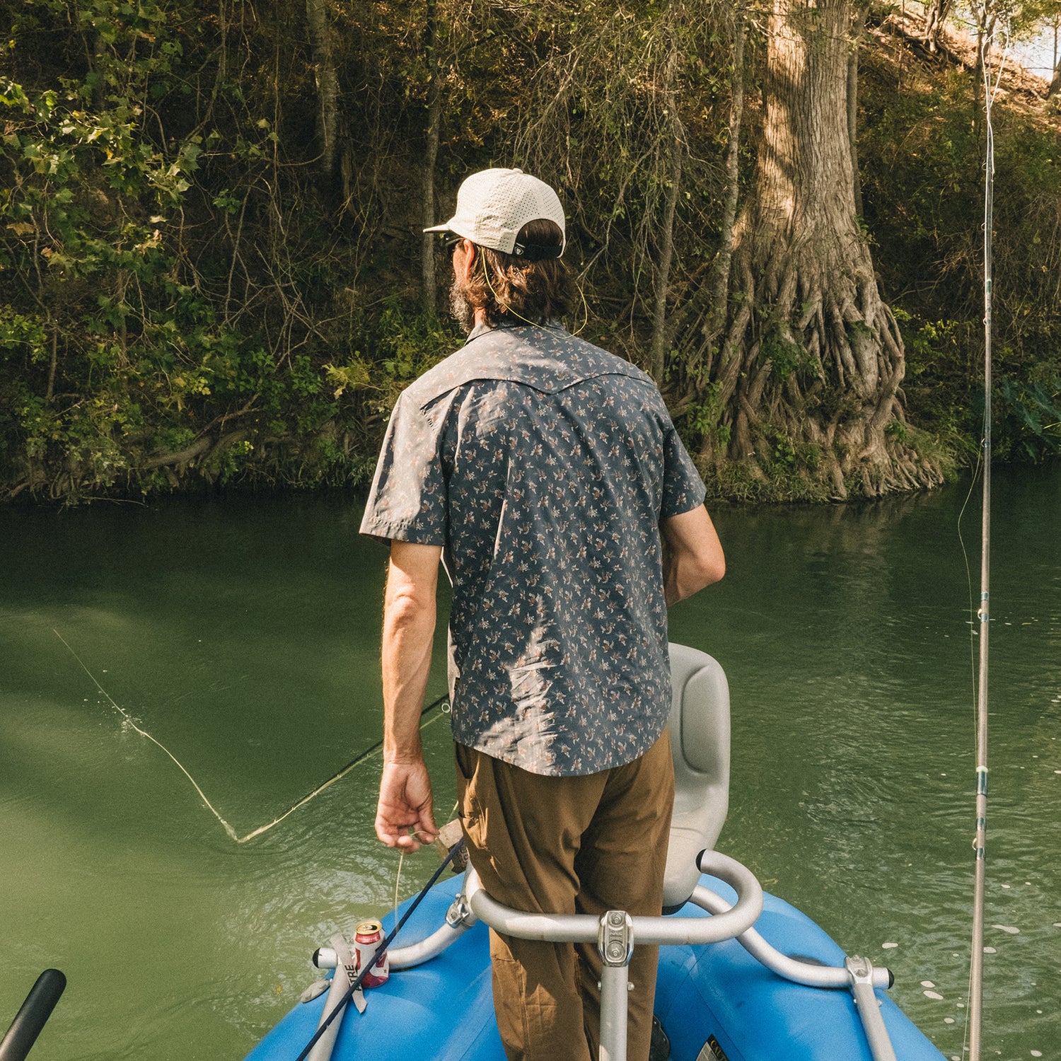 A man in a pearl snap shirt and cap stands at the edge of a blue inflatable boat, fishing on a calm river surrounded by lush greenery and tree roots along the riverbank. - View 8