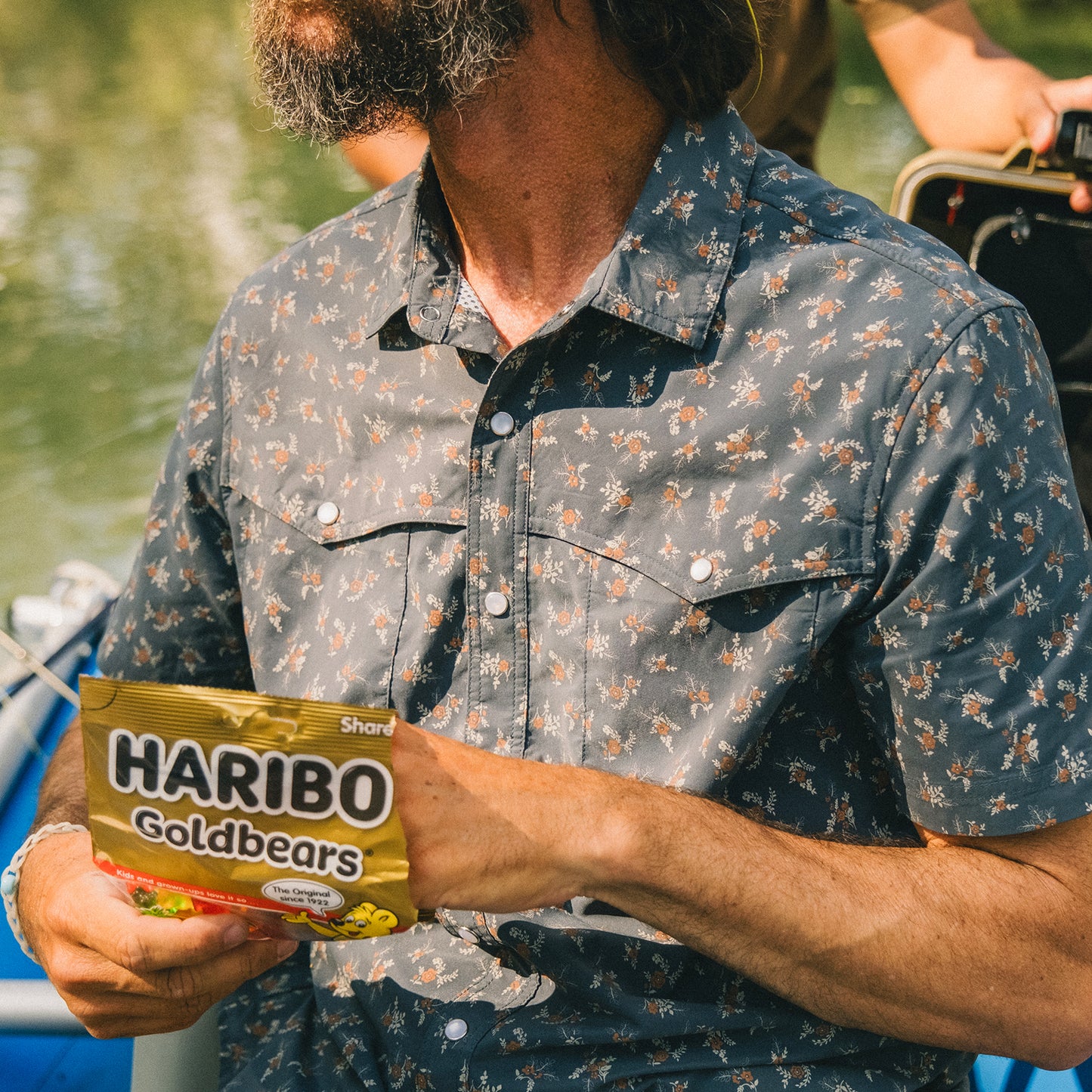 A bearded man in a pearl snap shirt holds a bag of HARIBO Goldbears gummy candy while sitting outdoors, possibly on a boat near water. - View 7
