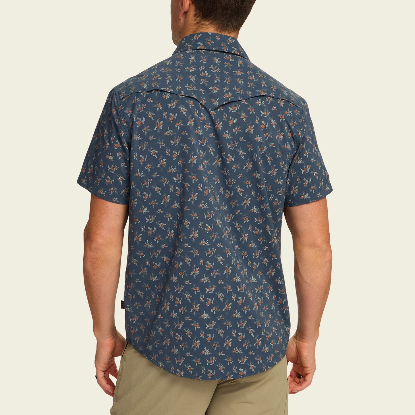 A man is standing with his back to the camera, wearing a blue technical utility shirt with a small floral pattern and beige shorts. The short-sleeve shirt features a vented rear yoke for added comfort. - View 5