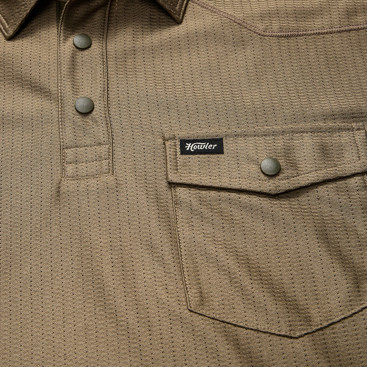 Close-up of a khaki textured shirt crafted from recycled polyester, featuring snap buttons, a flap chest pocket with a snap closure, and a small black Howler brand tag above the pocket. - View 4