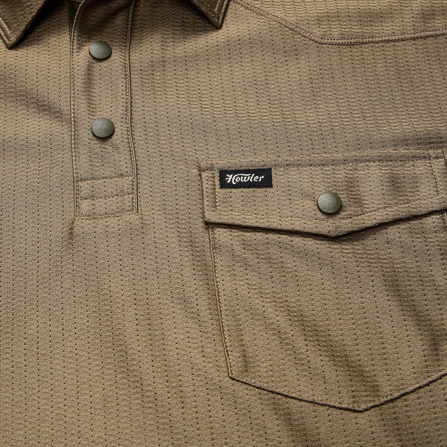 Close-up of a khaki textured shirt crafted from recycled polyester, featuring snap buttons, a flap chest pocket with a snap closure, and a small black Howler brand tag above the pocket. - View 4