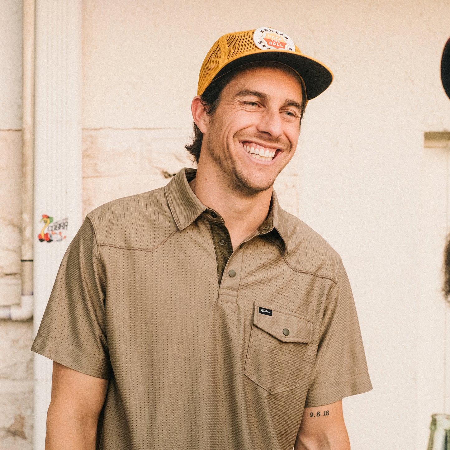 A smiling man in a tan tech polo and mustard yellow cap stands outdoors. He has a tattoo with numbers on his right forearm and is positioned in front of a light-colored wall with pipes and a sticker. - View 2