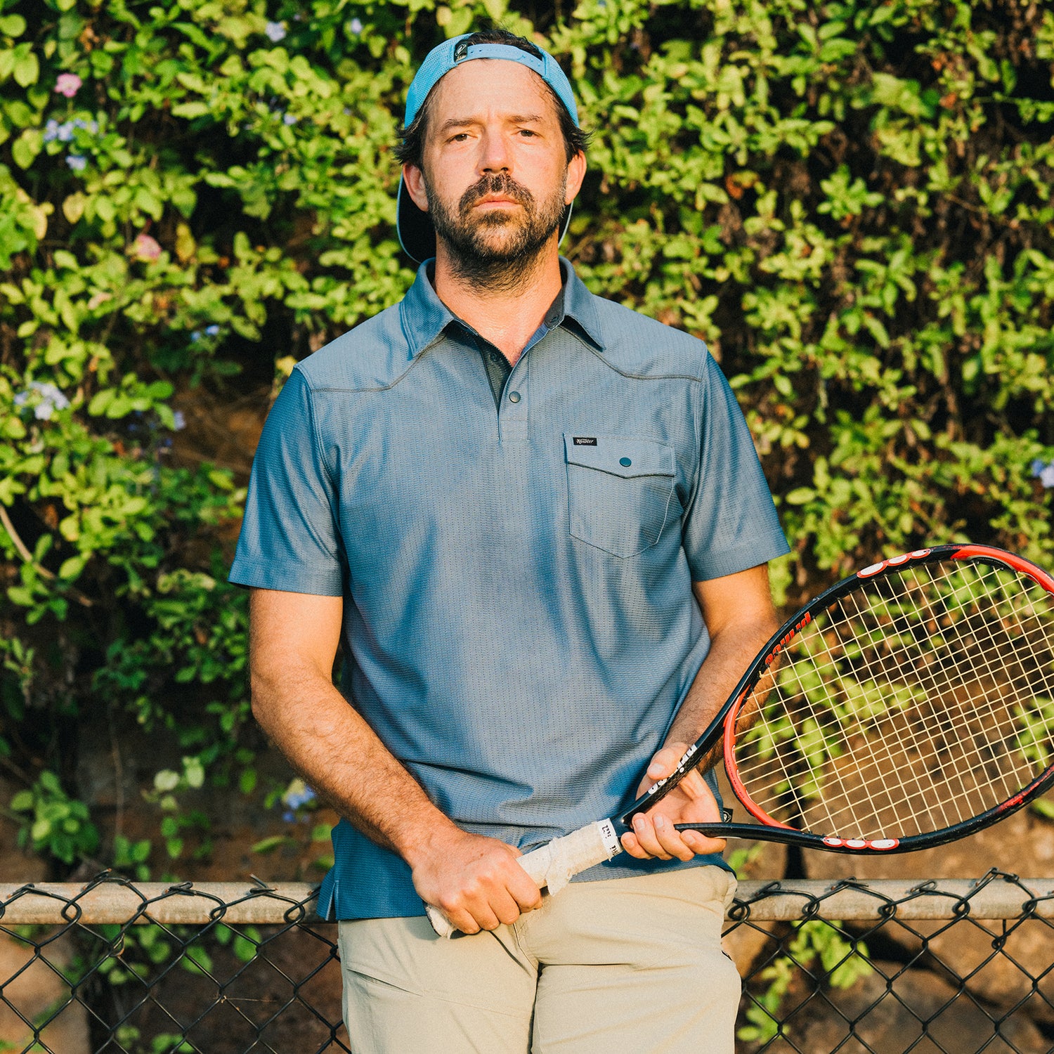A man in a blue Tech Polo made from breathable recycled polyester stands outdoors in front of leafy greenery, holding a tennis racket and leaning on a chain-link fence.