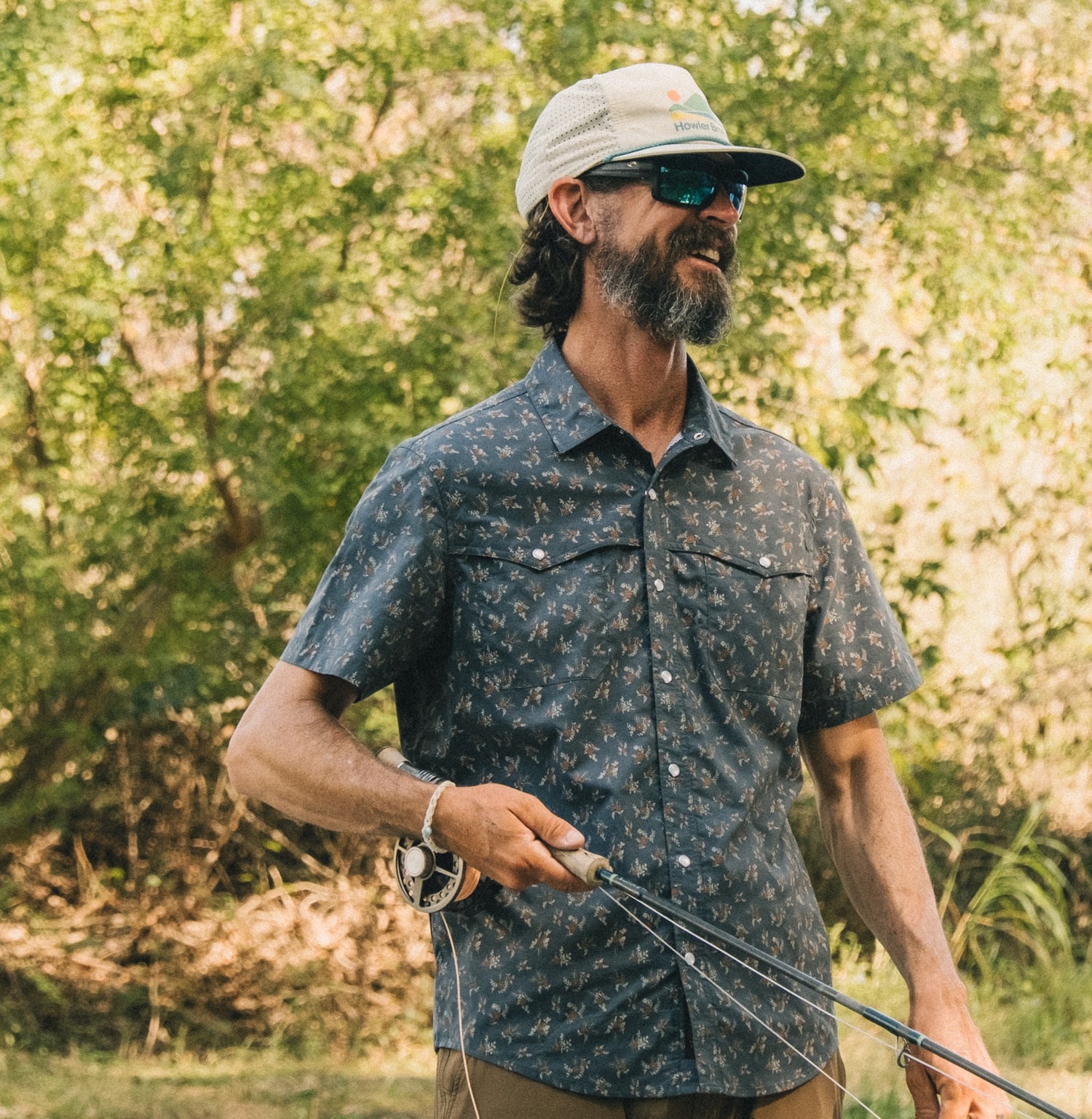 A man with a beard and sunglasses, wearing a patterned technical utility shirt with a vented rear yoke and a cap, stands outdoors holding a fishing rod, surrounded by green trees and sunlight.