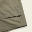 Close-up of olive green Quonset Camp Shorts with visible stitching, vintage-style buttons, and a small black and white Howler brand tag sewn near the pocket hem against a light background.