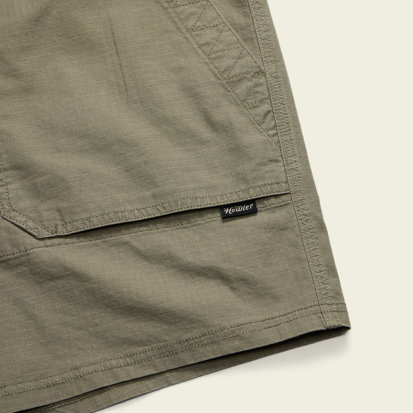 Close-up of olive green Quonset Camp Shorts with visible stitching, vintage-style buttons, and a small black and white Howler brand tag sewn near the pocket hem against a light background. - View 5