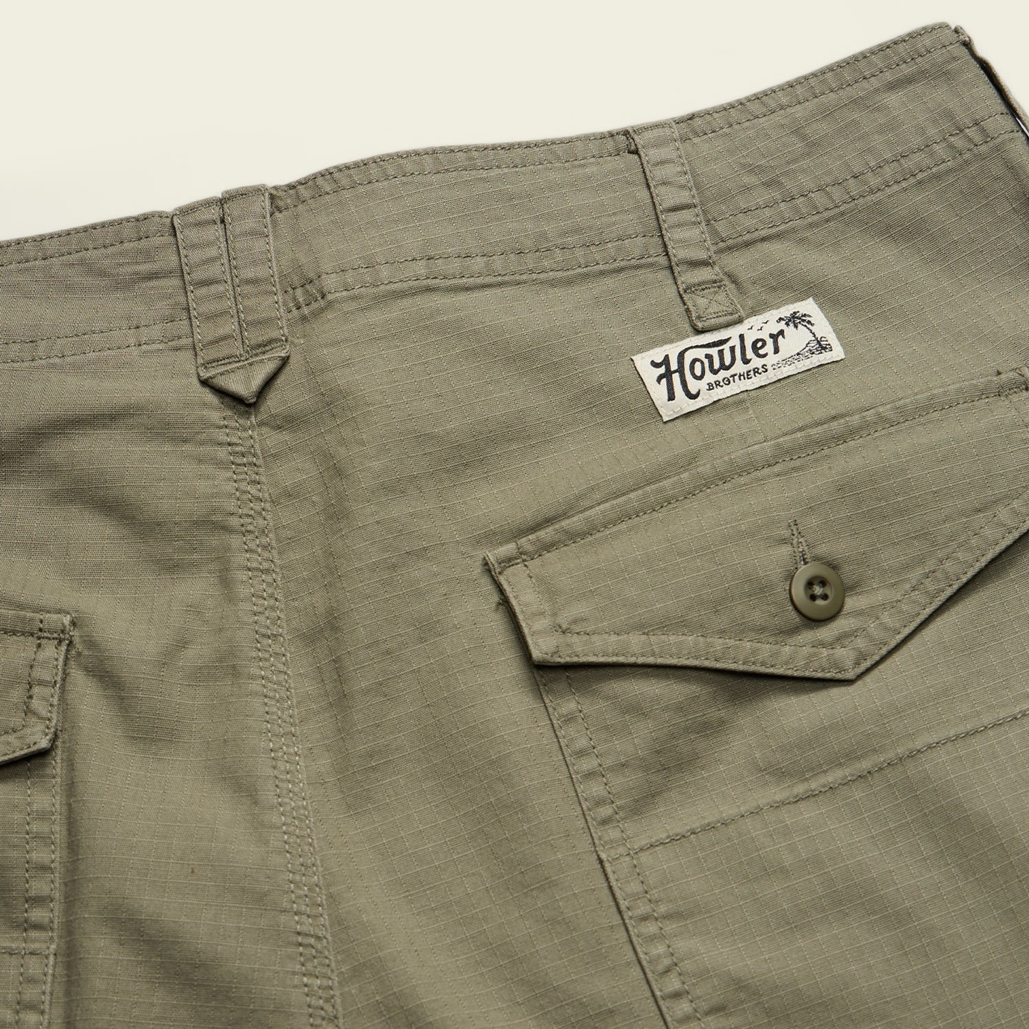 Close-up of olive green Quonset Camp Shorts crafted from ripstop cotton, featuring vintage-style buttons, belt loops, a flap pocket, and a Howler Brothers logo patch above the pocket. - View 6