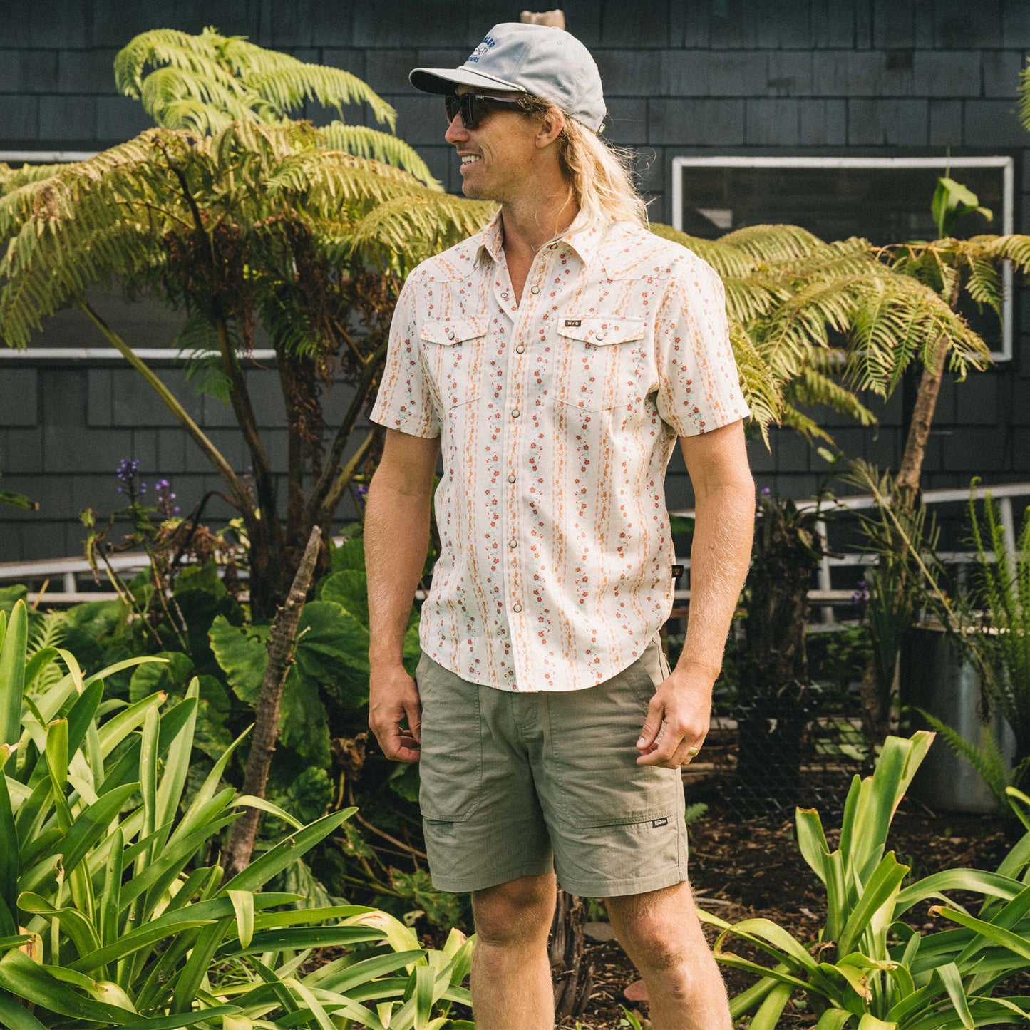 A man wearing a light patterned short-sleeve shirt, Quonset Camp Shorts with vintage-style buttons, and a gray cap stands smiling among green plants in front of a dark building with large windows. - View 2