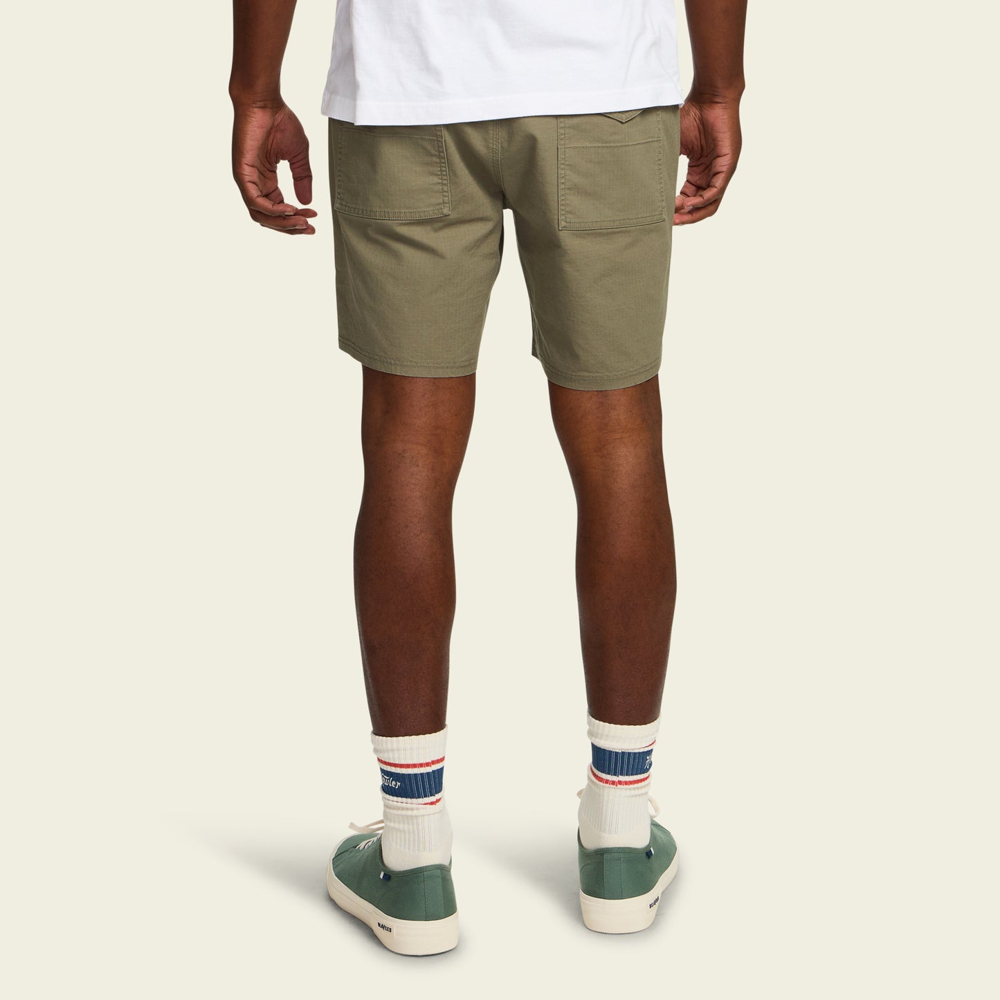 A person stands facing away, wearing Quonset Camp Shorts in olive green ripstop cotton, a white T-shirt, white socks with blue and red stripes, and green sneakers, set against a plain light background. - View 4
