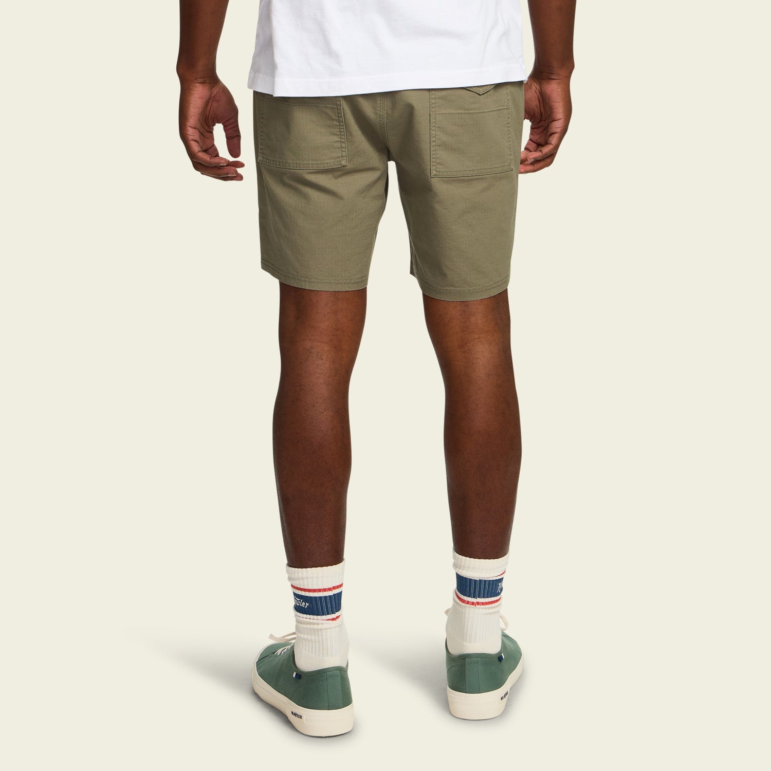 A person stands facing away, wearing Quonset Camp Shorts in olive green ripstop cotton, a white T-shirt, white socks with blue and red stripes, and green sneakers, set against a plain light background. - View 4