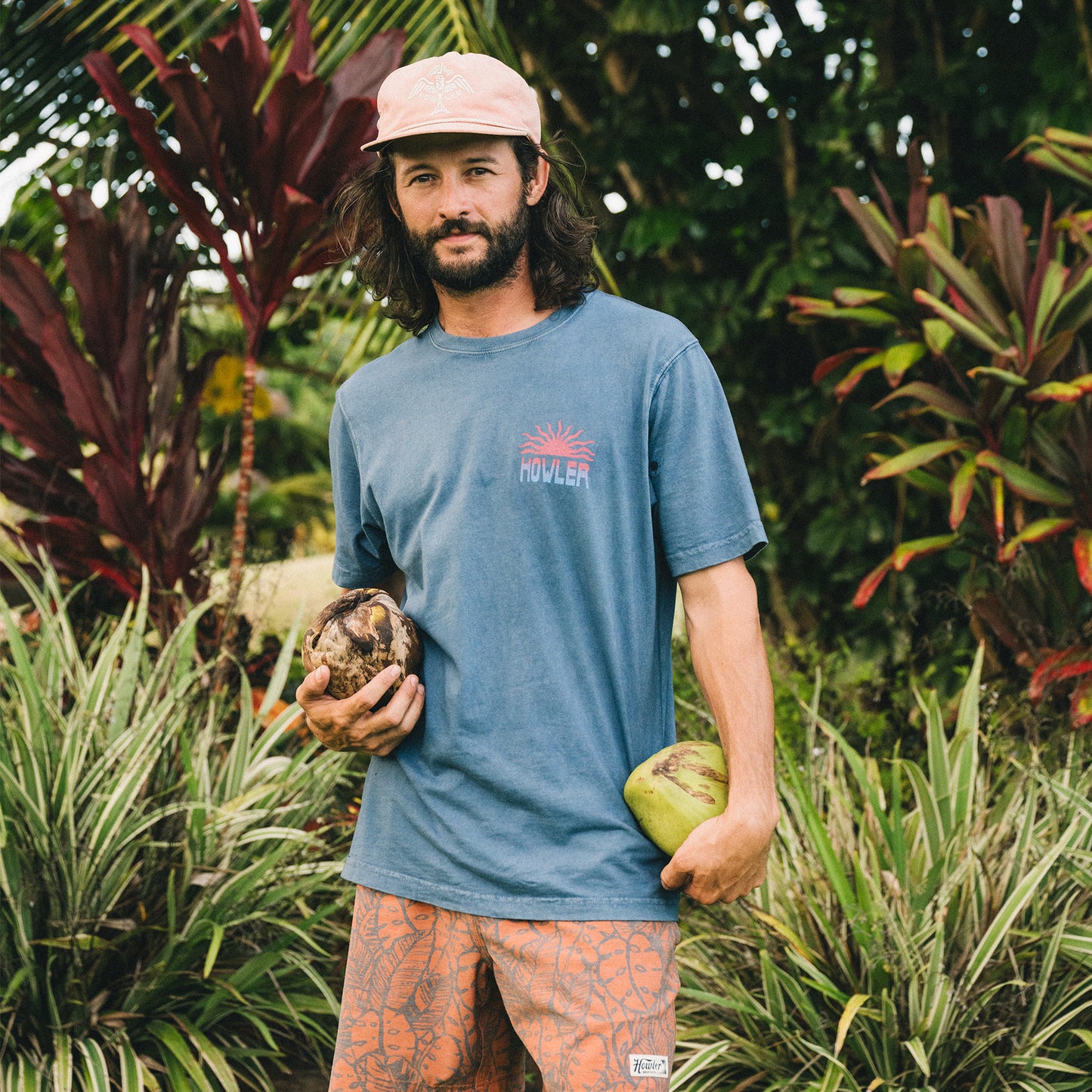 A man with long hair and a beard stands outdoors among tropical plants, wearing a vintage look blue cotton T-shirt, patterned shorts, and a pink cap. He holds two coconuts as he looks at the camera. - View 6