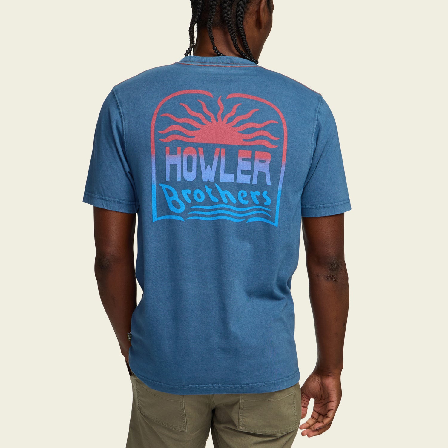 A person wearing a resin-washed blue cotton t-shirt with a red sun design and HOWLER Brothers printed on the back in red, white, and blue, paired with khaki pants. The vintage look is completed as the person is seen from behind. - View 5