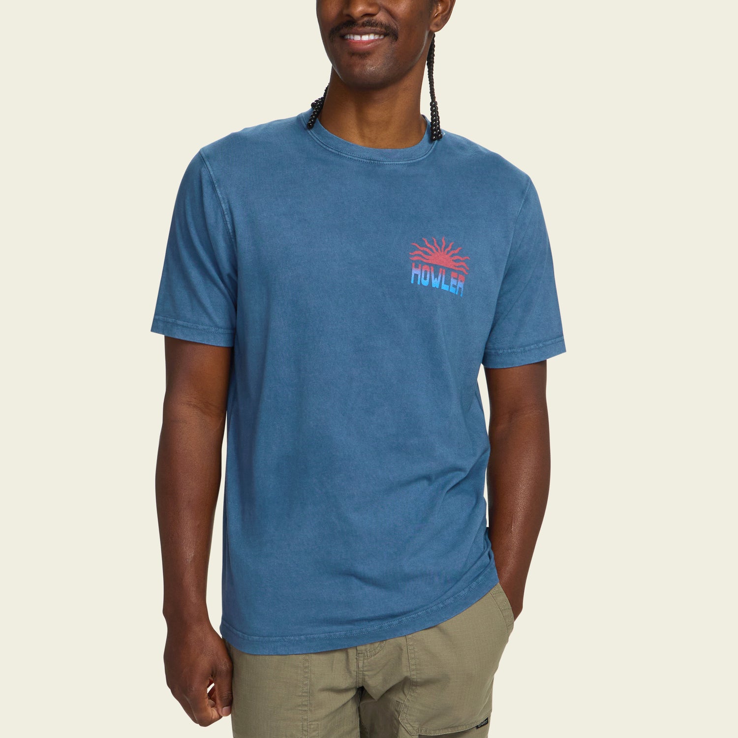 A man wearing a resin-washed blue short-sleeve cotton t-shirt with a small red and blue HOWLER graphic on the chest, and light green shorts, stands smiling against a plain light background for a vintage look. - View 4