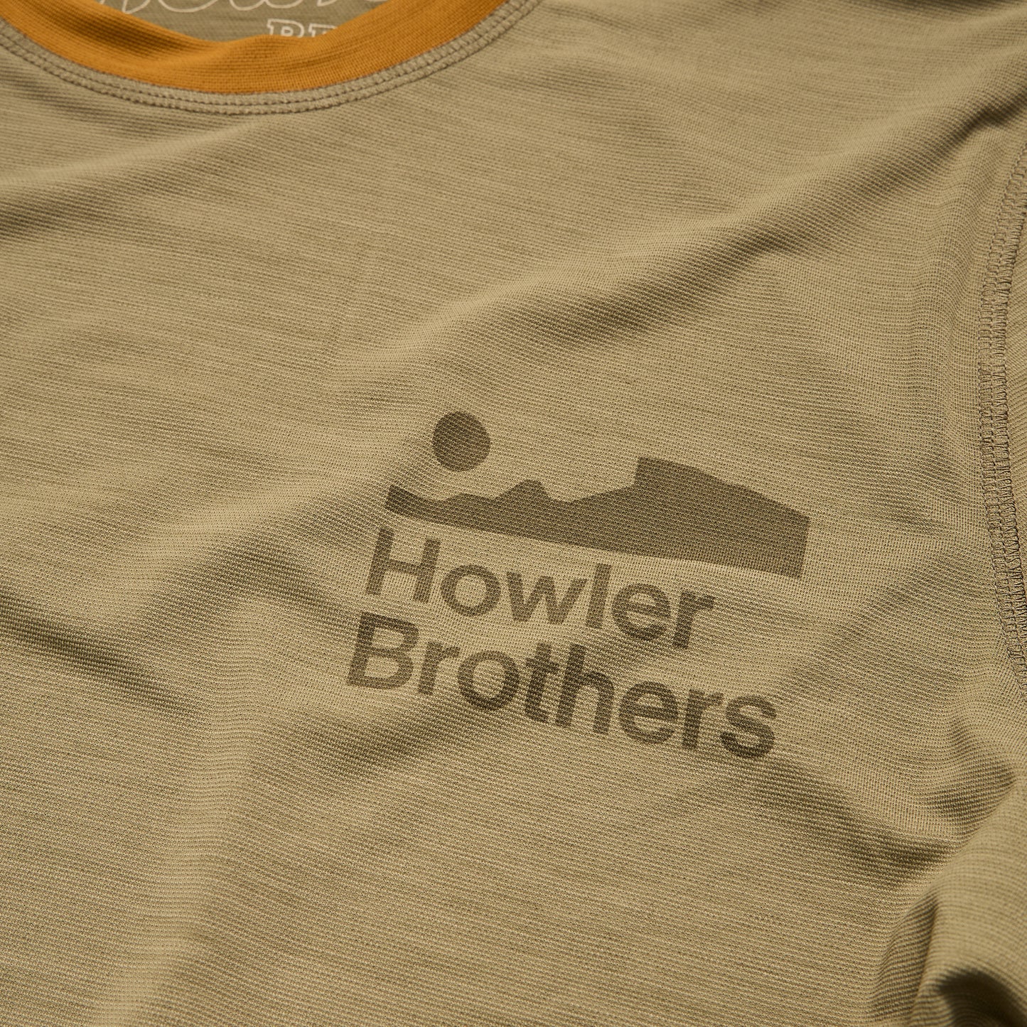 A close-up of a tan polyester spandex blend shirt with the Howler Brothers logo printed in dark brown, featuring stylized text below a minimalist landscape graphic with a sun and mountains. - View 5