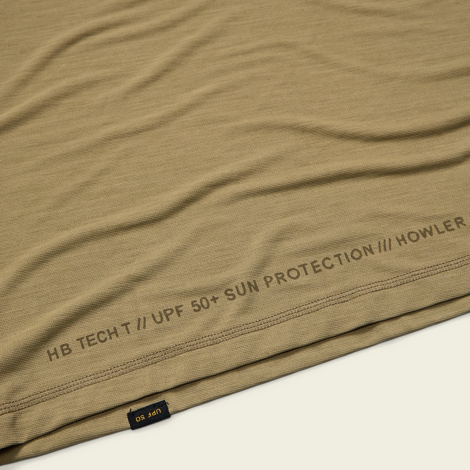 Close-up of a tan lightweight UPF shirt with HB TECH T // UPF 50+ SUN PROTECTION /// HOWLER printed near the hem and a small black and yellow UPF 50 tag on the seam. Made from a soft polyester spandex blend. - View 6