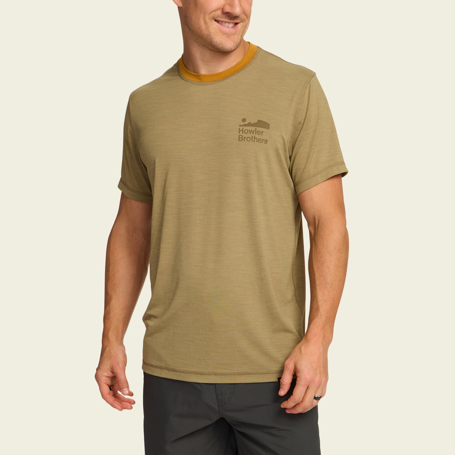A smiling man wears a light brown Howler Brothers shortsleeve tech shirt with a mustard-colored neckline and dark shorts, standing against a plain off-white background. - View 3