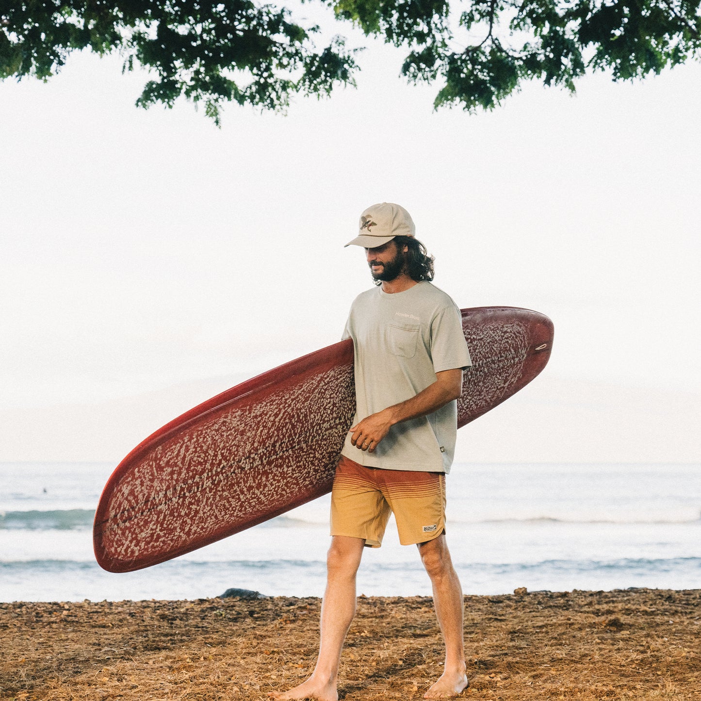 A man with long hair and a beard, wearing a beige cap, tan shorts, and a light green Howler blended t-shirt for maximum comfort, walks barefoot on a sandy beach carrying a red surfboard. The ocean and tree branches are in the background. - View 2