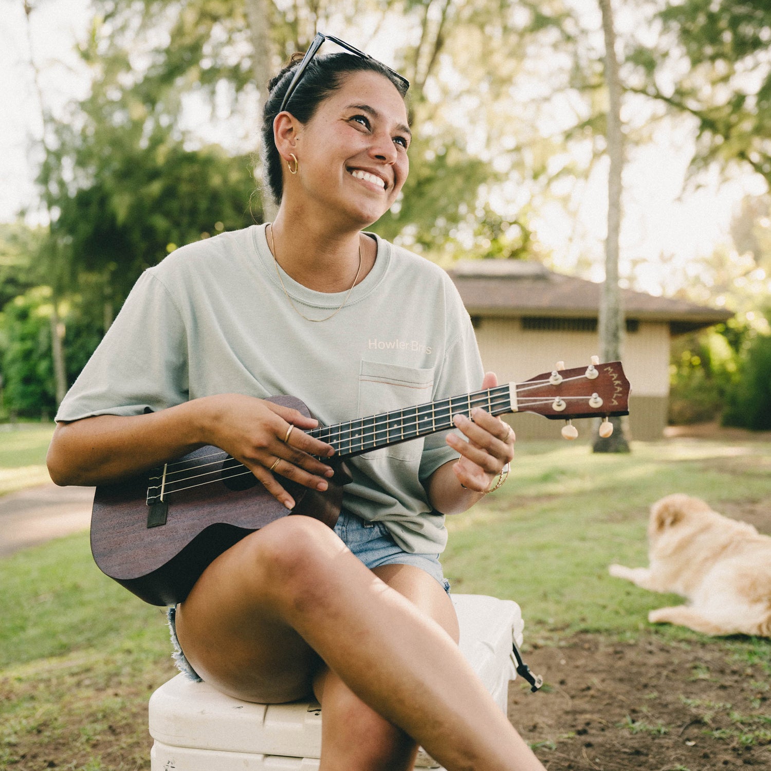 A smiling woman sits outdoors on a cooler playing a ukulele. She wears a Howler blended t-shirt and shorts, enjoying maximum comfort. Sunglasses rest on her head, with trees, a building, and a dog lying on the grass nearby. - View 6