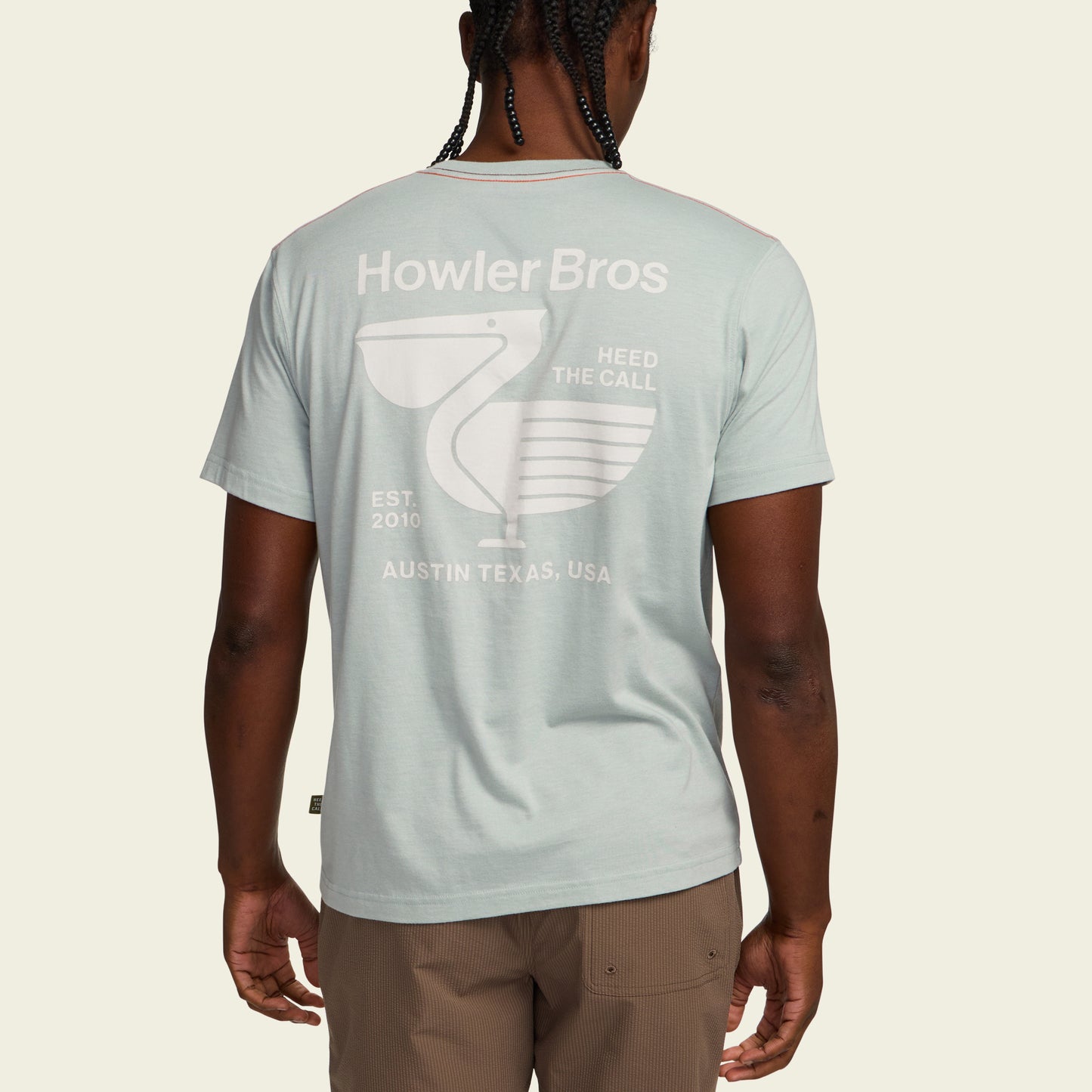 A person wearing a light blue Howler blended t-shirt featuring a pelican graphic and text, Heed the Call, Est. 2010, Austin Texas, USA, viewed from the back. Made from a soft cotton-poly blend for maximum comfort. - View 5