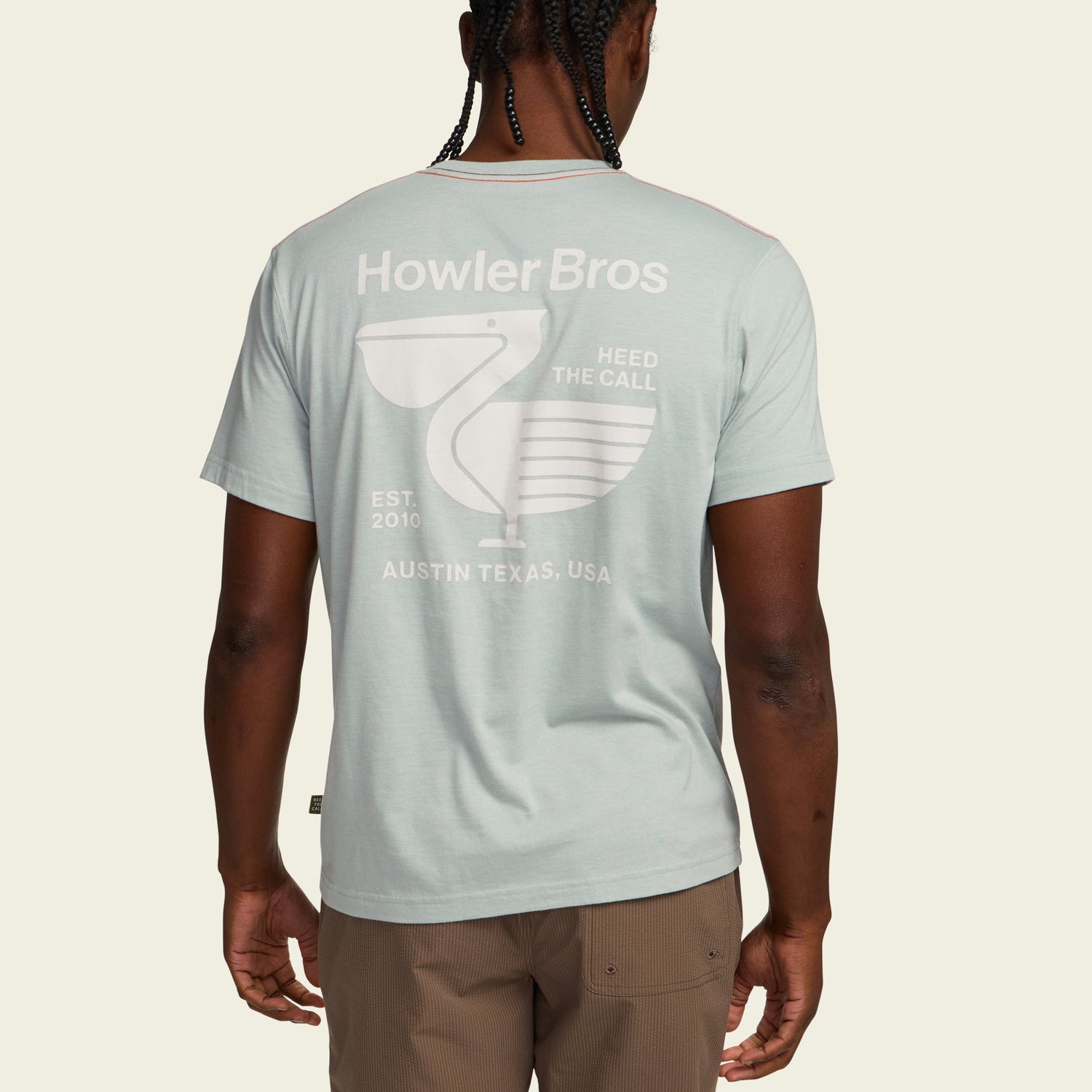 A person wearing a light blue Howler blended t-shirt featuring a pelican graphic and text, Heed the Call, Est. 2010, Austin Texas, USA, viewed from the back. Made from a soft cotton-poly blend for maximum comfort. - View 5