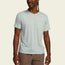 A man wearing a light green Howler blended t-shirt made from a soft cotton-poly blend with a chest pocket and Howler Bros text, paired with brown shorts, stands against a plain light background for maximum comfort.