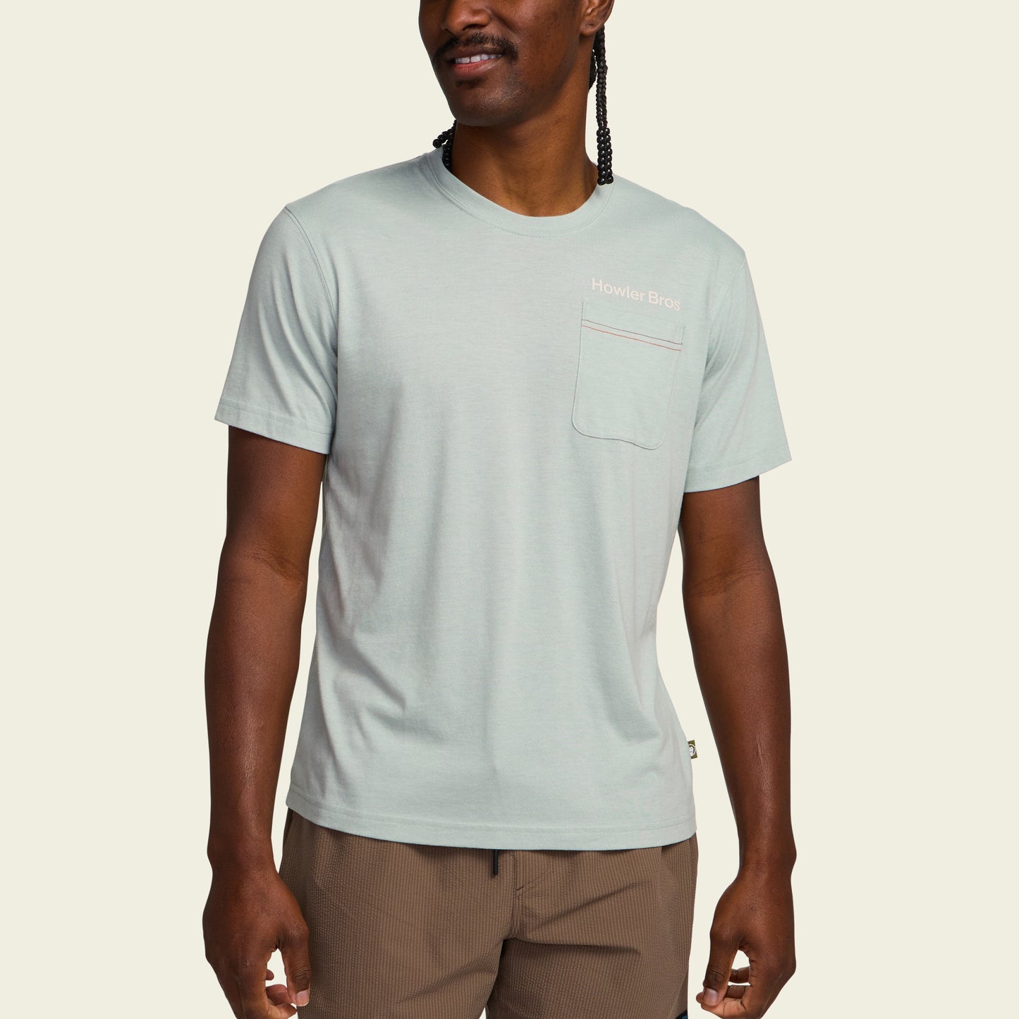 A man wearing a light green Howler blended t-shirt made from a soft cotton-poly blend with a chest pocket and Howler Bros text, paired with brown shorts, stands against a plain light background for maximum comfort. - View 4