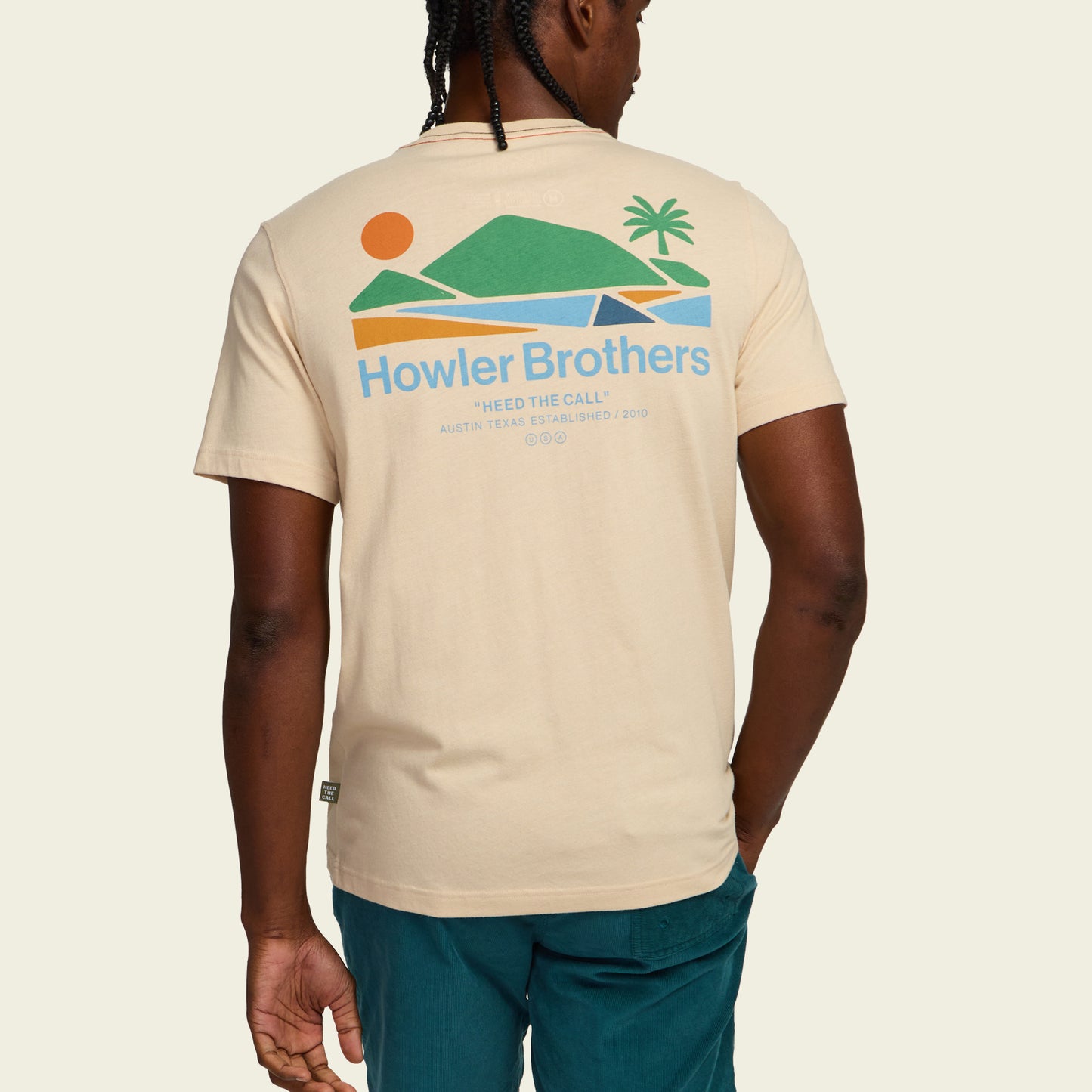 A man seen from behind wears a beige Howler blended t-shirt featuring a colorful graphic of a sunset, mountains, water, and palm tree, with the text Howler Brothers and Heed the Call, crafted for maximum comfort. - View 5