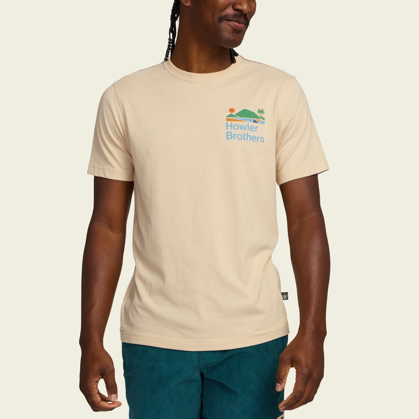 A person wearing a beige Howler blended t-shirt made from a soft cotton-poly blend for maximum comfort, featuring a small sun, wave, and palm tree graphic on the chest, paired with teal shorts against a plain background. - View 4