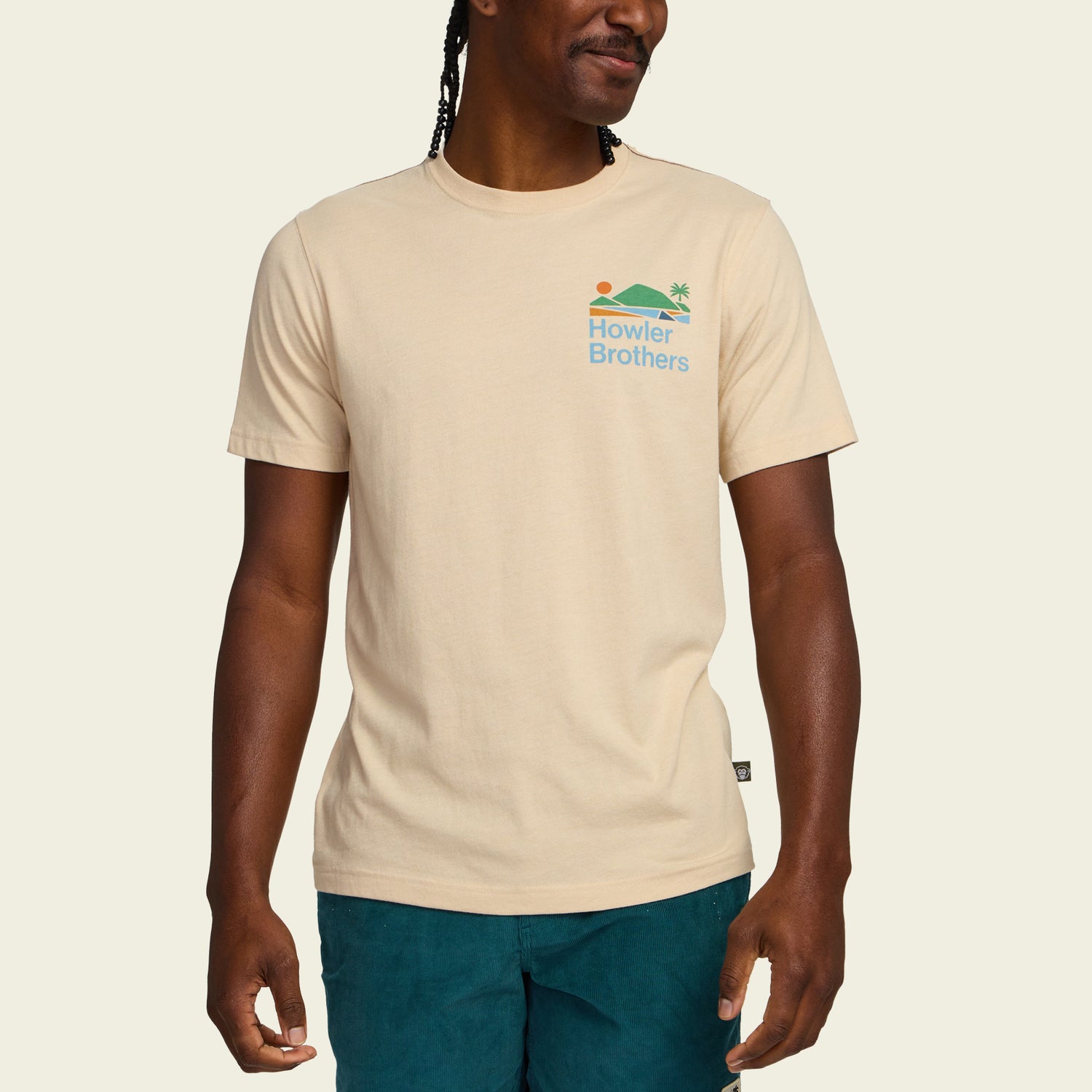 A person wearing a beige Howler blended t-shirt made from a soft cotton-poly blend for maximum comfort, featuring a small sun, wave, and palm tree graphic on the chest, paired with teal shorts against a plain background. - View 4