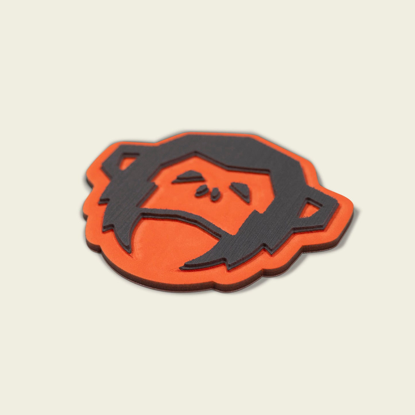A flat, orange and black object shaped like a cartoon monkeys face with a sad expression, crafted from high-density foam as the El Mono Hook Pad, shown on a plain light background. - View 4