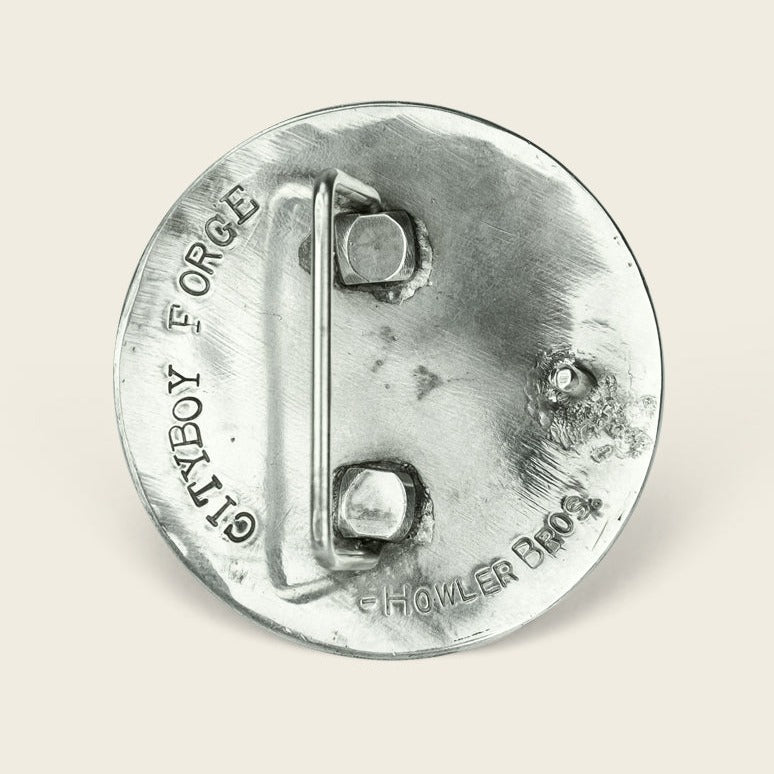 A round, silver metallic object with a handle and two bolts, engraved with GRITTY BY FORCE and Howler Brothers on its surface, resembles a sterling silver agave buckle, set against a plain light background. - View 3