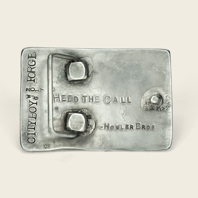 A rectangular metal object with two raised square pegs and engraved text reading HEED THE CALL - HOWLER BROS and CITYBOY FORGE along the side. This custom coordinates buckle features a slightly textured and tarnished surface. - View 3