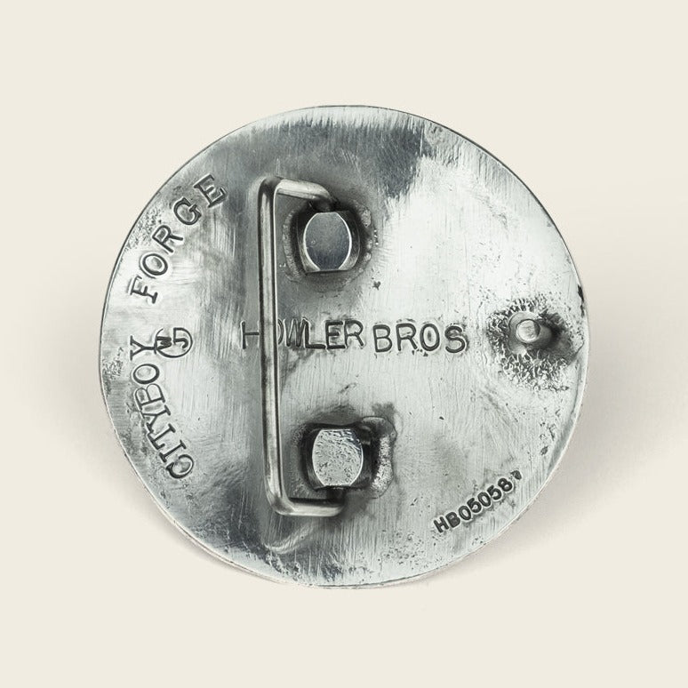 A round metal object with raised text reading Cityboy Forge and HOWLER BROS, reminiscent of an El Gallo buckle, featuring two metal loops and a small stamped code HB05038 on a plain background. - View 3