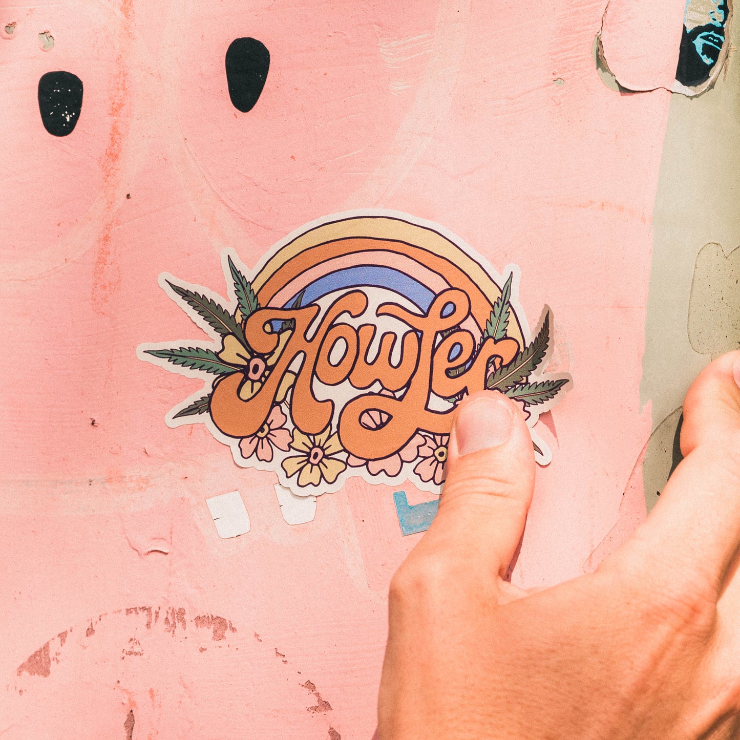 A hand holds a Howler Brothers sticker on a pink surface. The heavy duty sticker features the word Howler in retro script, framed by a rainbow arch, leaves, and flowers. - View 2