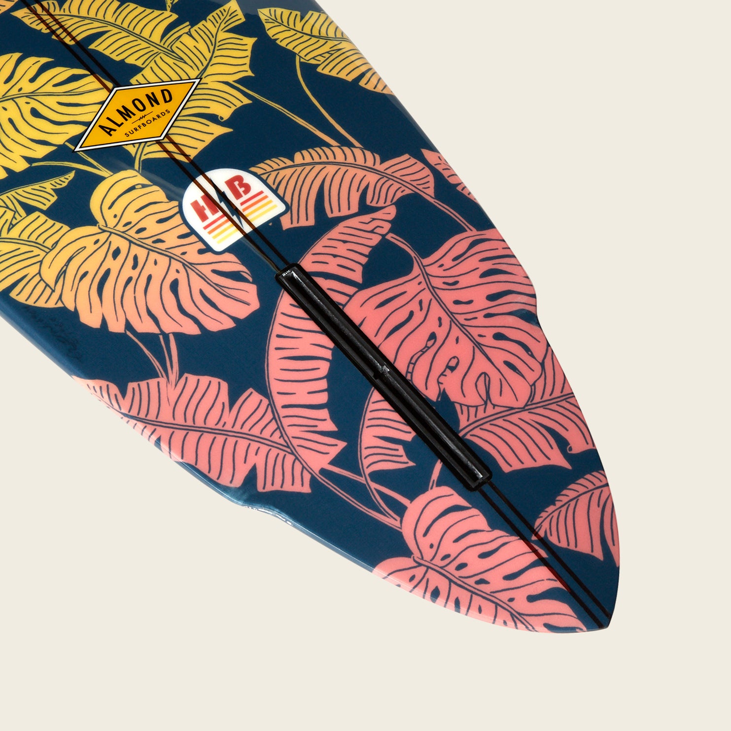 The tip of a surfboard decorated with pink and yellow tropical leaf patterns on a dark blue background, featuring brand stickers including one that says ALMOND from Almond Surfboards. - View 2