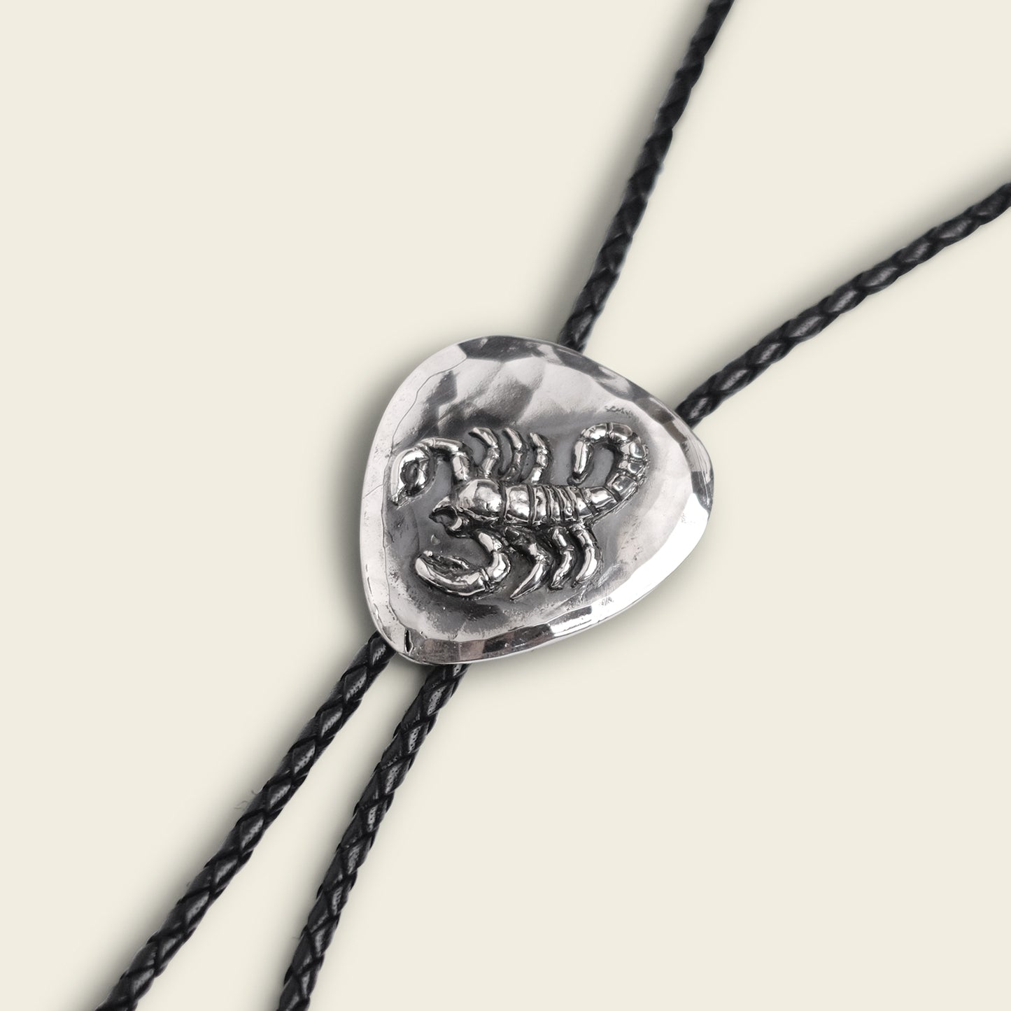A silver El Alacrán Bolo Tie slide with a raised scorpion design is threaded onto two black braided leather cords, set against a light background—an eye-catching handmade bolo tie by Cityboy Forge. - View 2
