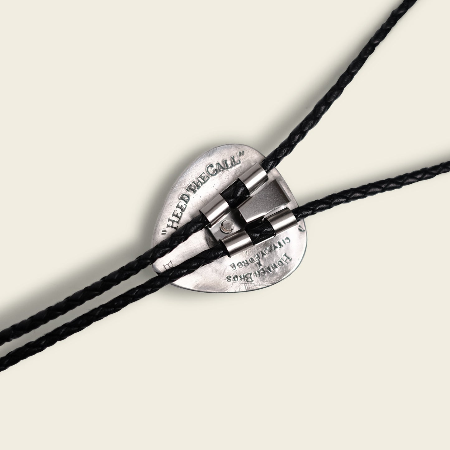 A close-up of a silver heart-shaped clasp with engraved text, securing two black braided leather cords—a handmade bolo tie by Cityboy Forge—against a light background. - View 3