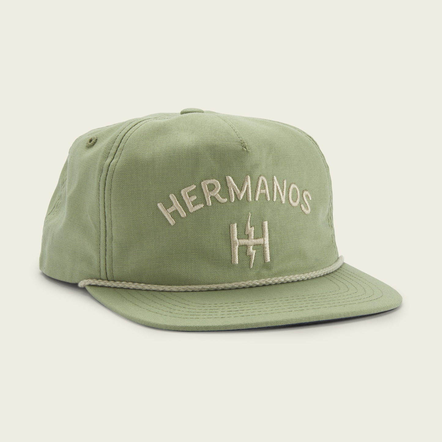 A light green snapback baseball cap with the word HERMANOS and a stylized H logo embroidered in white on the front, featuring a custom fit, flat brim design, and a braided rope detail above the brim. - View 2