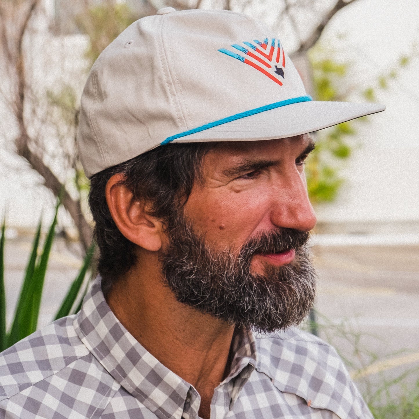 A man with a thick beard and mustache wears an unstructured snapback featuring a dark under brim and adjustable snap back closure, paired with a plaid shirt, standing outdoors amid greenery and a blurred background. - View 3